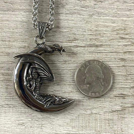 Silver dragon pendant on a chain next to a quarter for scale on a wooden surface