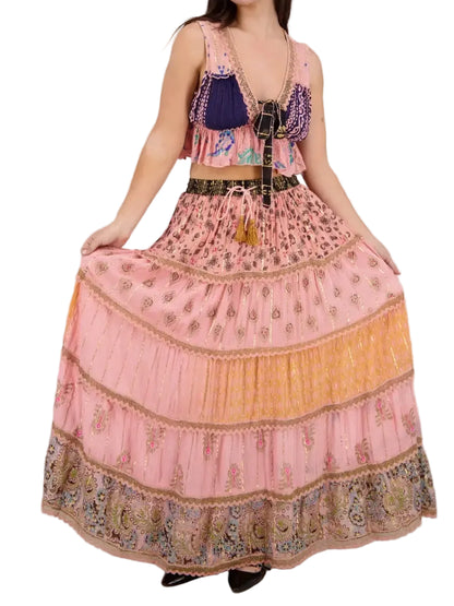A woman modeling a bohemian-style two-piece outfit featuring a tiered pink maxi skirt with gold and orange patterns, paired with a matching crop top that has a navy blue and purple embroidered bodice.
