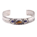 An antique-style silver cuff bracelet featuring a central oval brown tiger's eye stone and two small round blue gemstone accents, set against a plain white background.