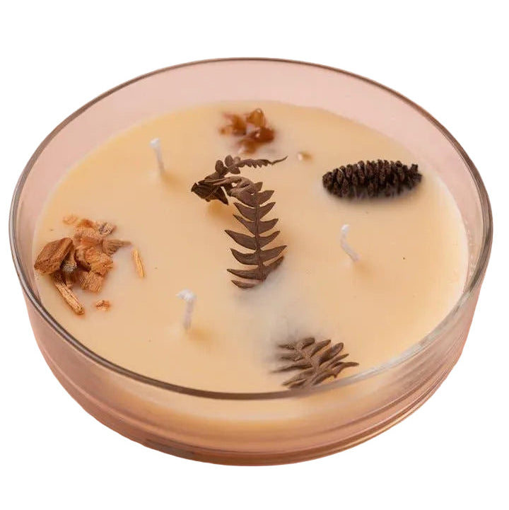 A clear glass jar candle filled with creamy beige wax and decorated with dried botanical elements including a fern leaf, pinecone, and small brown flowers, set against a solid pale pink background.