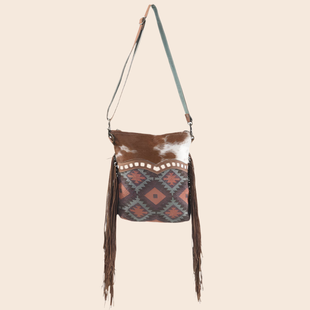 Brown and white cowhide bag with patterned flap and fringes on a white background