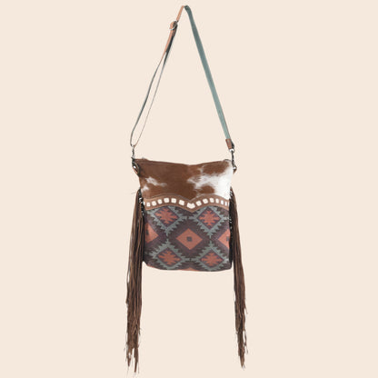 Brown and white cowhide bag with patterned flap and fringes on a white background