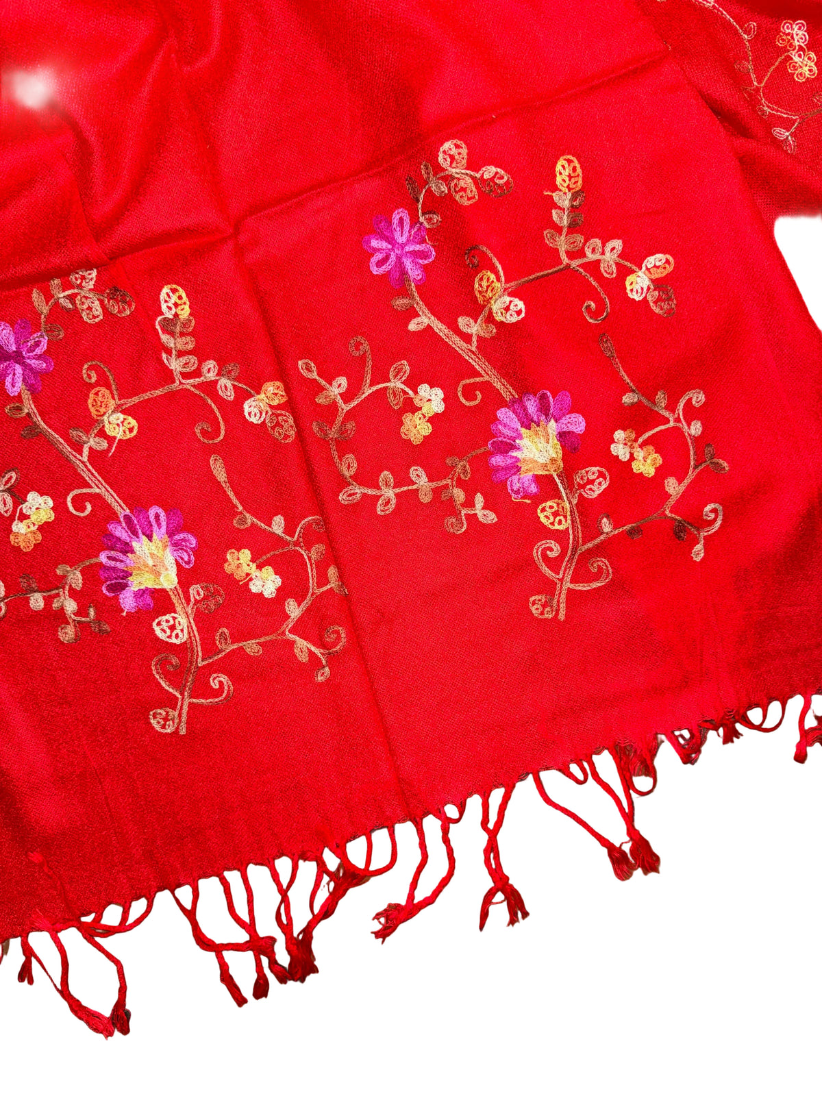 A close-up of a bright red fabric with embroidered floral and vine motifs in pink, pink, and gold, finished with red fringe tassels on a white background